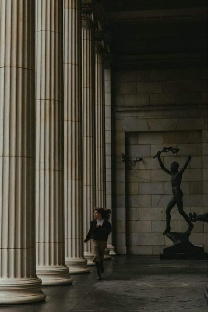 Person walking by classical columns and a statue in a dimly lit historic building corridor.