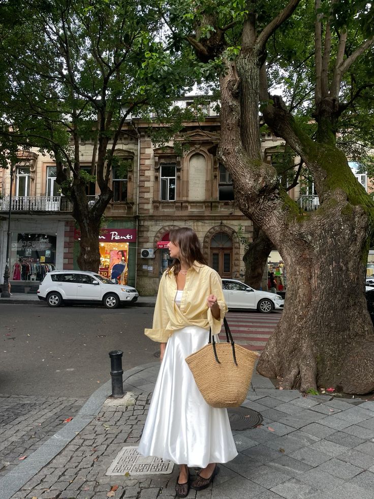 Woman in stylish outfit with straw bag stands under trees on an urban street, showcasing a blend of nature and city life.