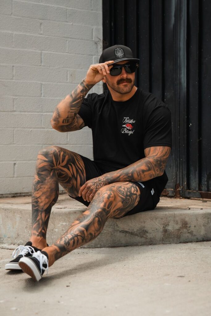 Tattooed man in black attire sitting on concrete step, adjusting sunglasses, showcasing stylish urban fashion.