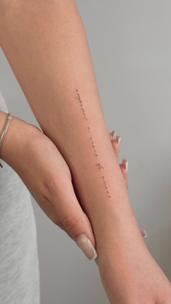 Minimalist script tattoo on forearm, person with neutral nails and bracelet, against a plain background. Tattoos for Women