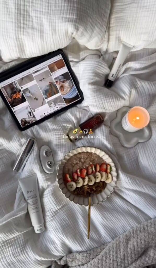 Cozy morning setup with oatmeal, skincare products, a lit candle, and a tablet on a soft bedspread.