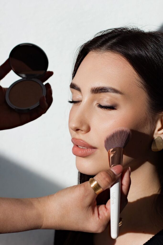 Woman getting makeup applied with brush and bronzer in natural light.