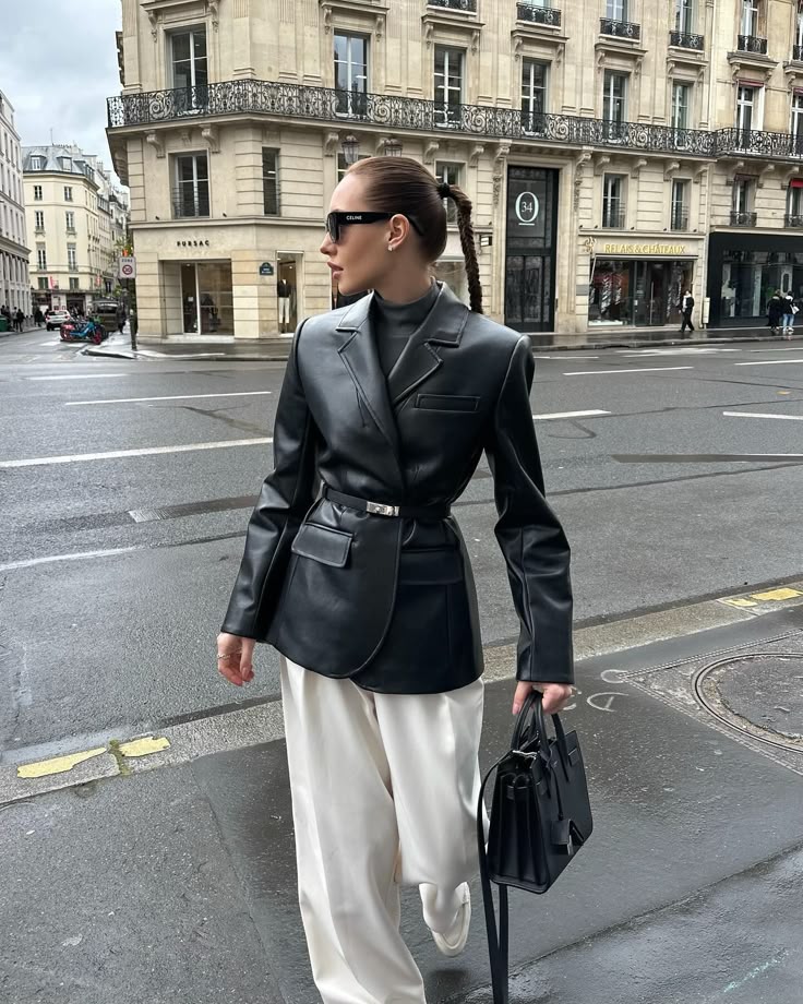 Fashionable woman in black leather jacket and white pants, walking in a city street, holding a black handbag.