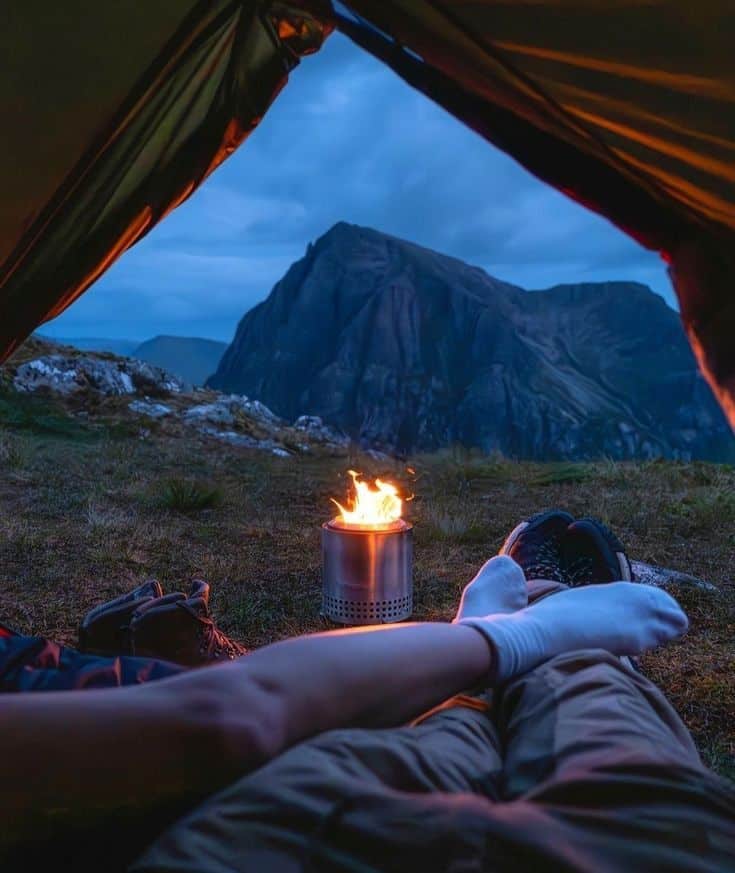 Cozy tent camping with view of mountains and campfire, relaxing outdoors under twilight sky.
