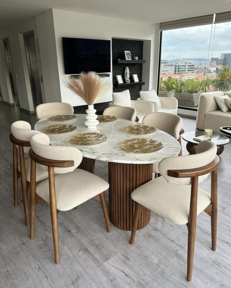 Modern dining room with a round marble table, wooden chairs, and city view through large windows.