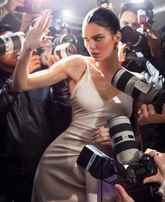 Glamorous woman in elegant dress surrounded by photographers, striking a pose in the spotlight.