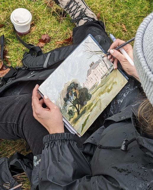 Person sitting on grass, sketching a landscape painting of a building and trees, with coffee cup nearby.