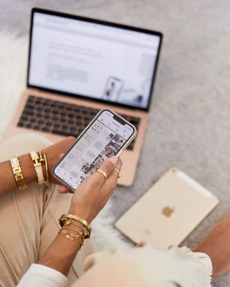 Person browsing Instagram on smartphone, with laptop and tablet nearby on a carpeted floor.