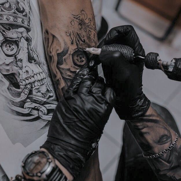 Tattoo artist creates detailed skull design on arm using needle and ink, with reference image on paper nearby.