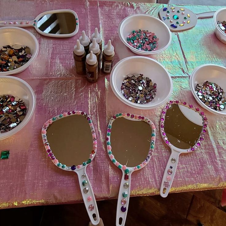 Decorated hand mirrors with colorful gems on a pink table, alongside bowls of rhinestones and glue bottles.