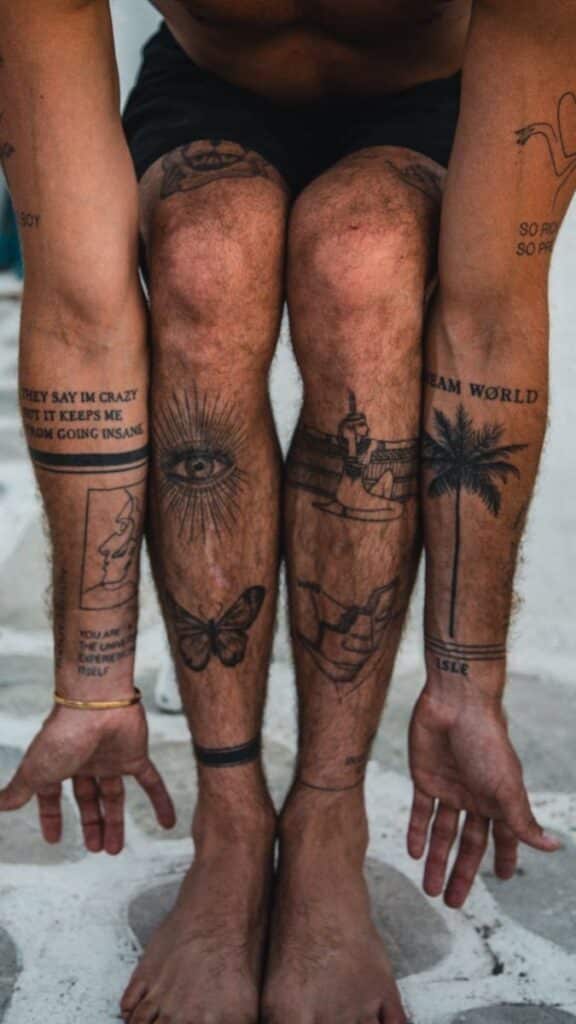 Person's tattooed legs and forearms with various designs like an eye, butterfly, and palm tree, against stone background.