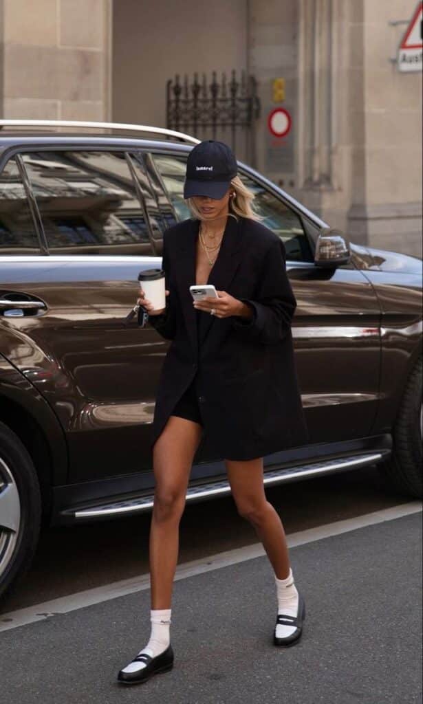 Fashionable woman in oversized blazer and cap walking with coffee, phone in hand, beside a parked car. Urban street style.