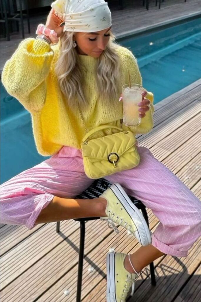 Fashionable woman in yellow sweater and pink pants by pool, holding drink. Stylish outfit with matching bag and shoes.