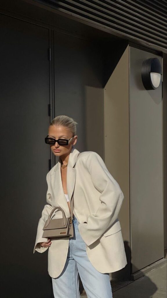 Fashionable woman in a white blazer and jeans with a small handbag, wearing sunglasses, standing outside in sunlight.