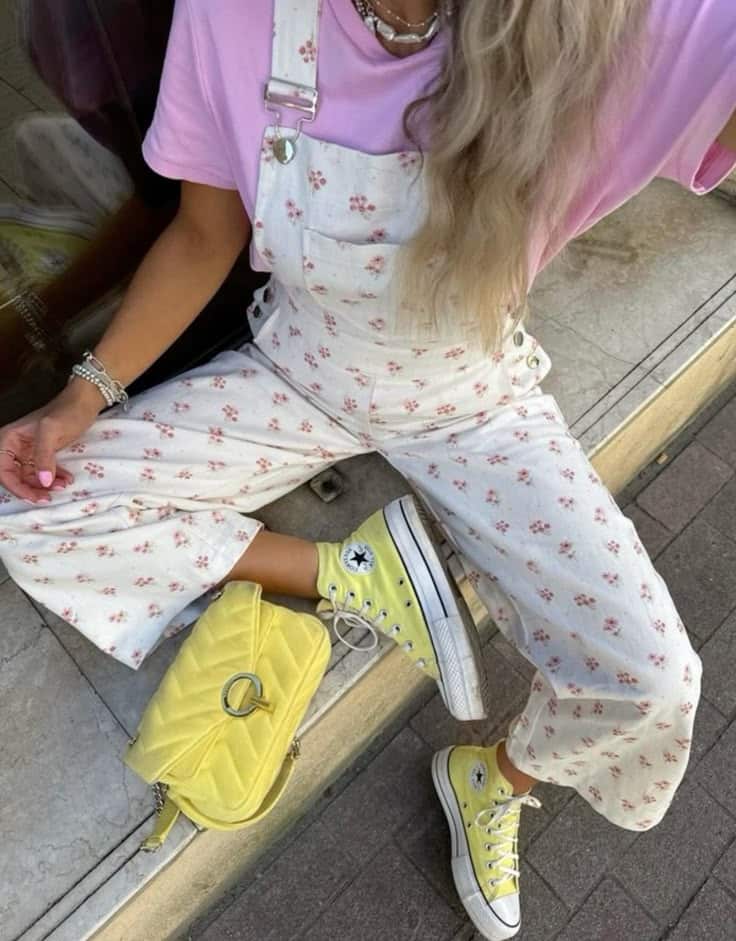 Woman in floral overalls and pink top with yellow Converse and bag, sitting on stone steps. Casual and stylish look.