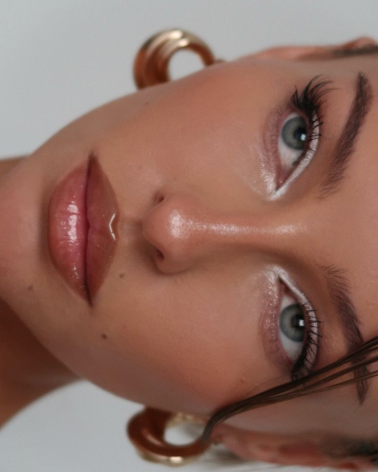Close-up of a woman's face with glossy lips and silver eye makeup, wearing bold gold earrings.