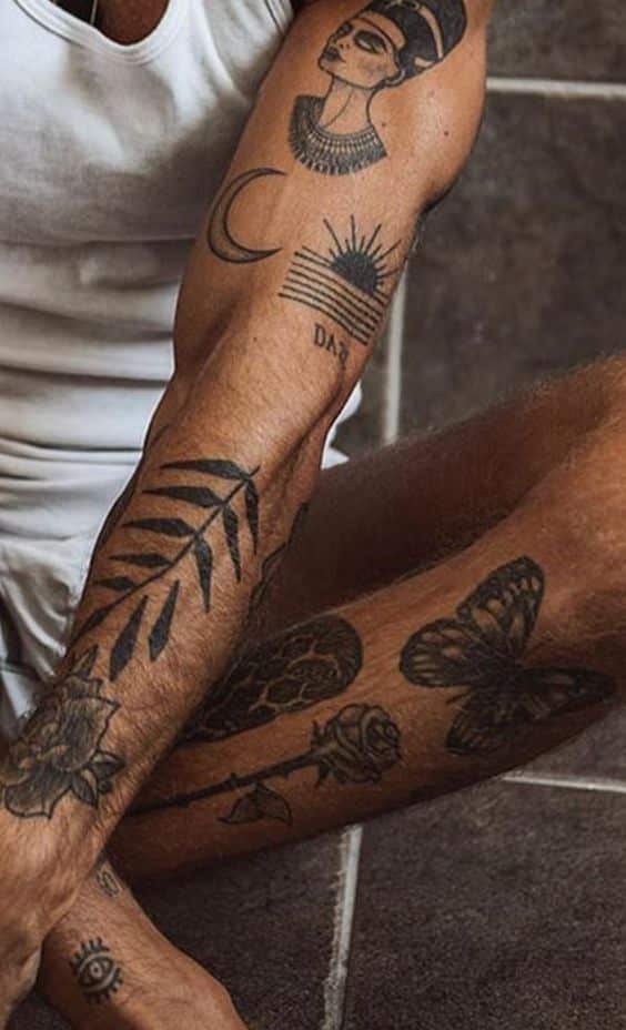 Man with arm and leg tattoos featuring a face, crescent moon, leaf, and butterfly, wearing a white tank top, sitting indoors. Men Tattoo Ideas