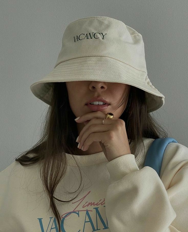 Stylish woman in a beige bucket hat and sweater, featuring VACANCY embroidery, posing with hand near face.