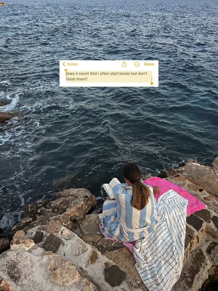 Person in striped robe reading by the sea on rocky shore with a note about not finishing books, reflecting tranquility.