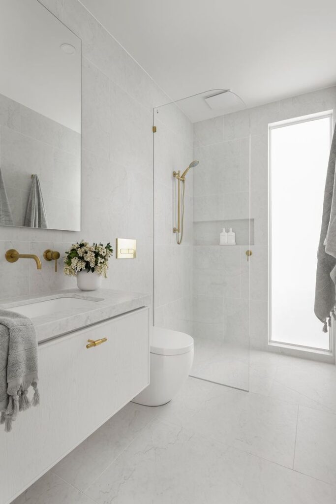 Minimalist white bathroom with gold fixtures, white vanity, flowers, and a glass shower partition.