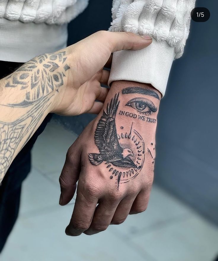 Hand tattoo featuring an eye, bird, and In God We Trust text, with intricate black ink design.