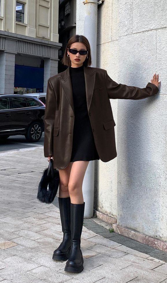 Stylish woman in oversized blazer, black dress, and boots, standing on urban street, exuding confidence and elegance.
