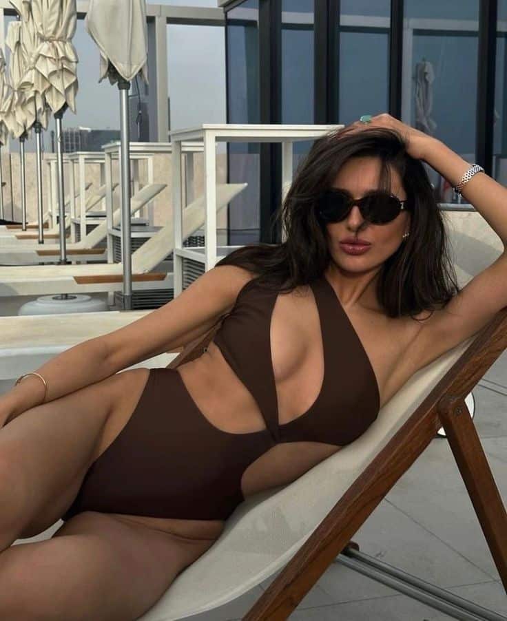 Woman relaxing in a stylish cutout swimsuit on a poolside lounger, wearing sunglasses.