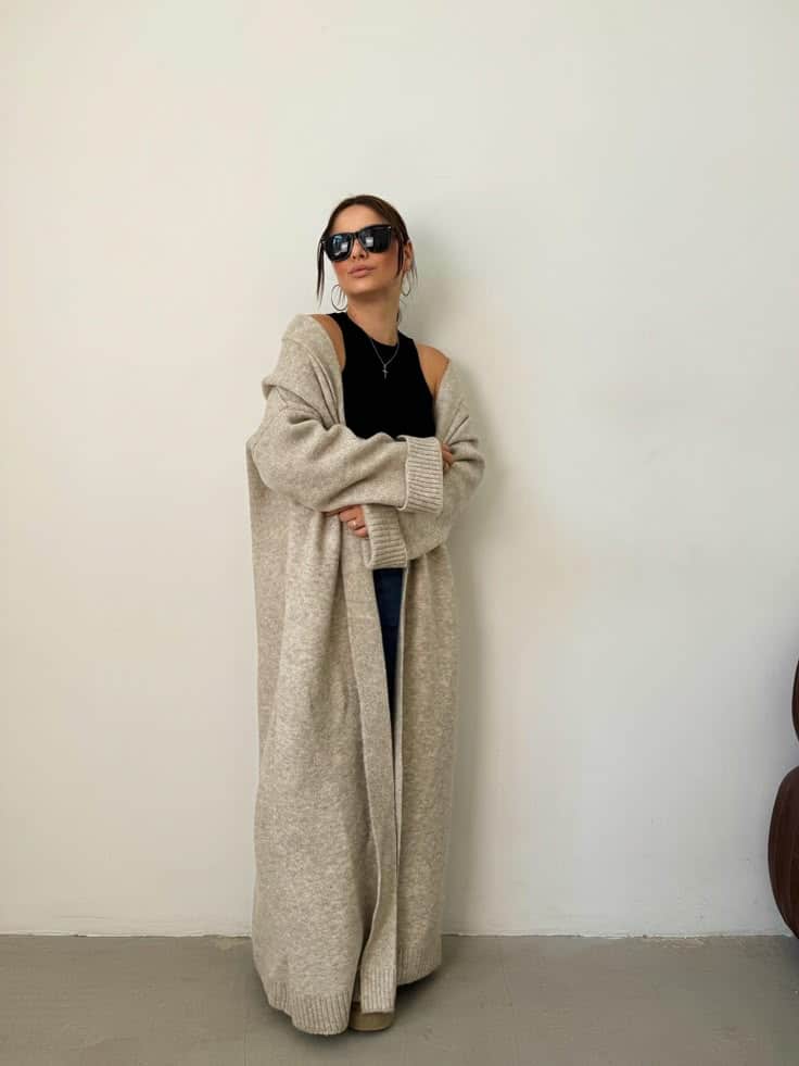 Woman in sunglasses wears a long beige cardigan and black top, posing confidently against a plain background. Fashion style.