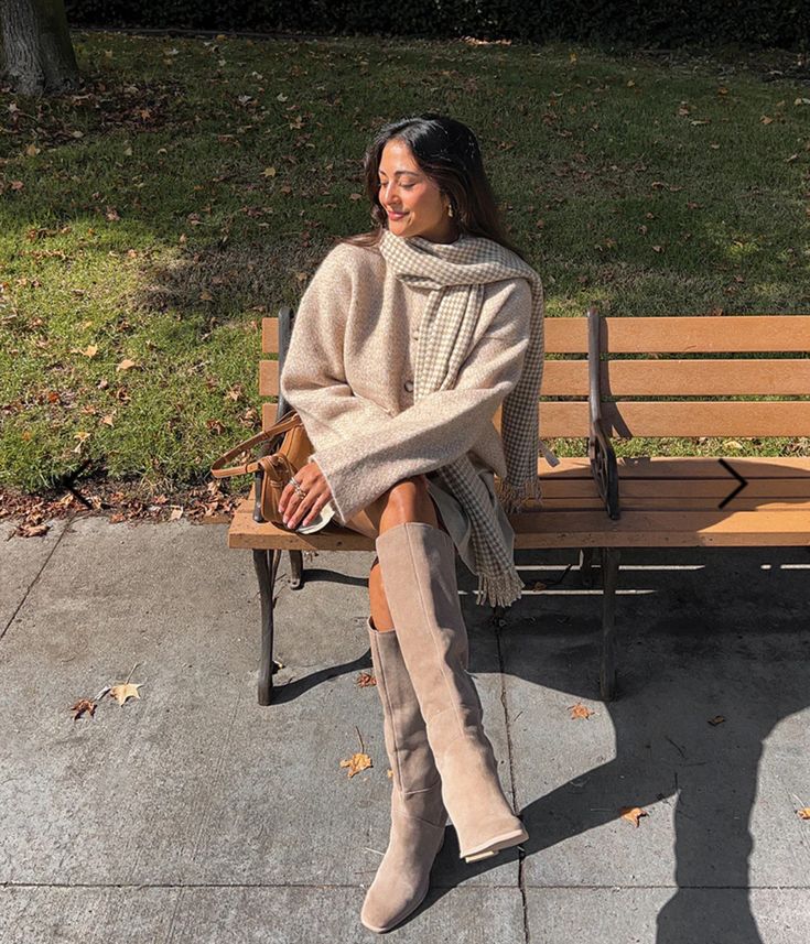 Woman in beige coat and scarf sits on park bench, basking in autumn sunlight.