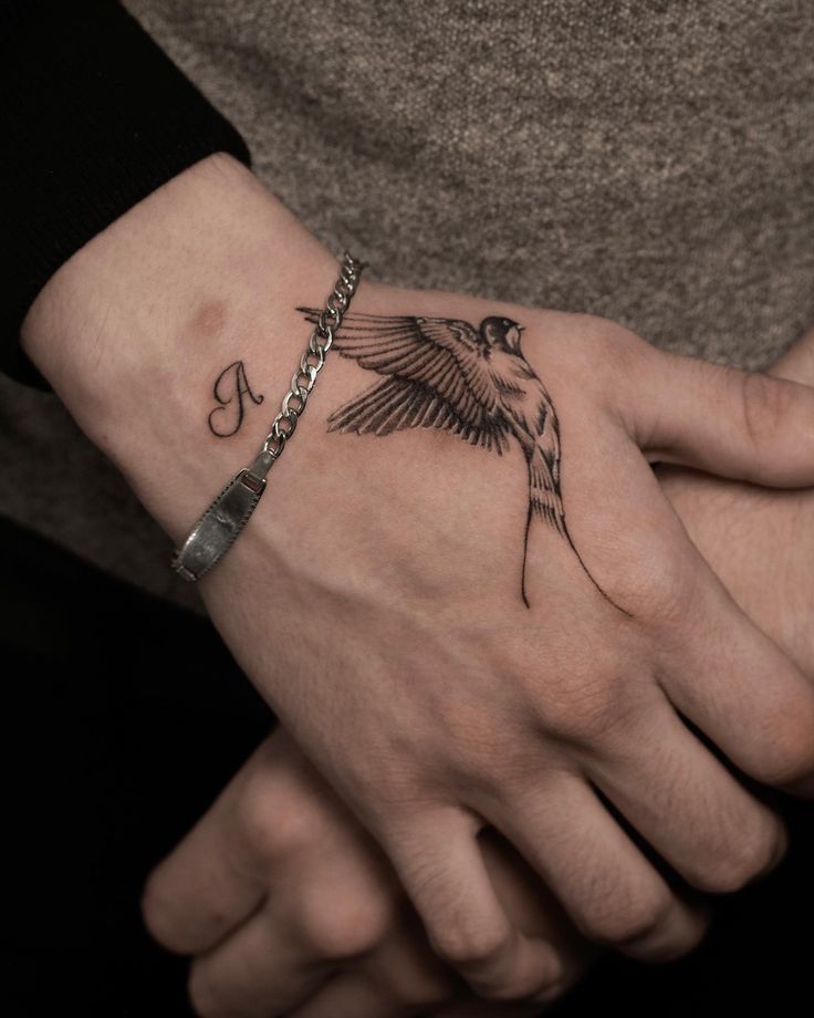 Hand with bird and letter A tattoos wearing a silver bracelet, close-up view.