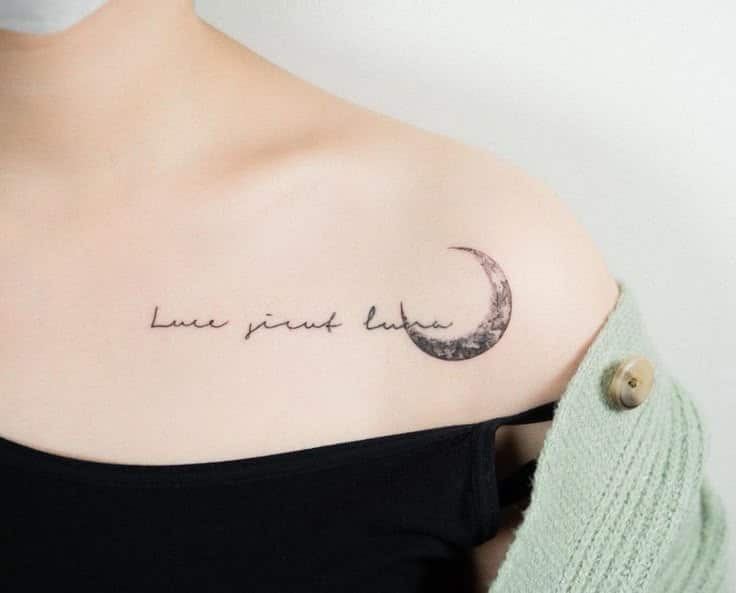 Minimalist shoulder tattoo with crescent moon and cursive script, worn by individual in black top and green sweater.