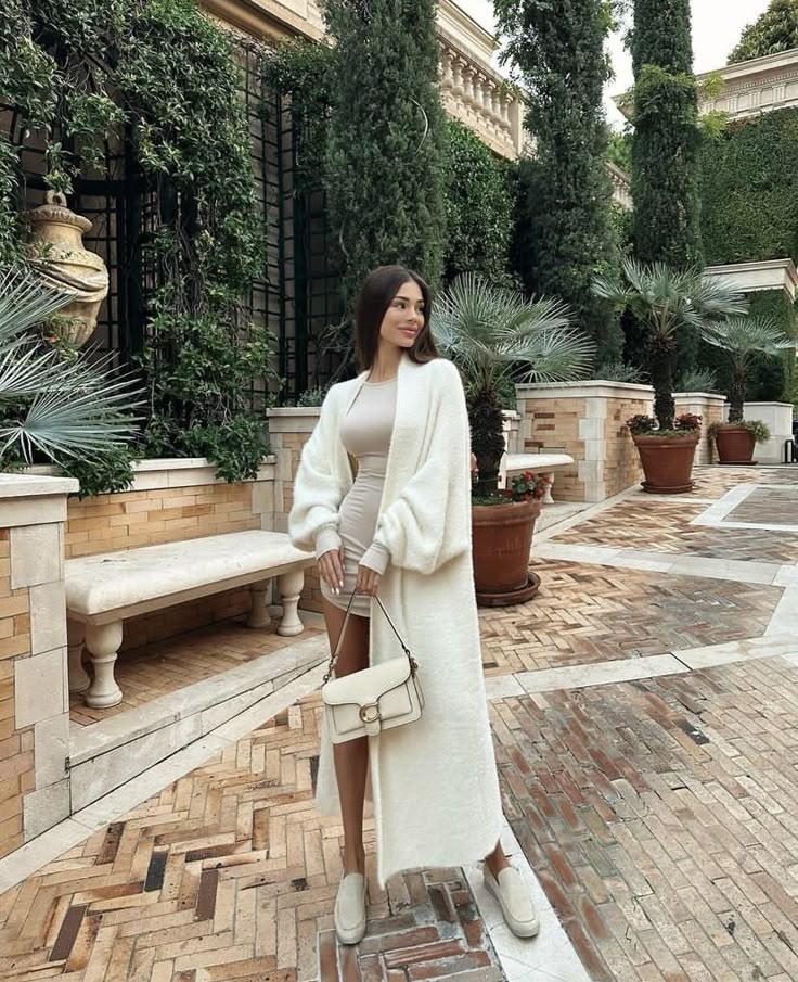 Woman in elegant outfit with white coat and purse in a scenic garden setting.