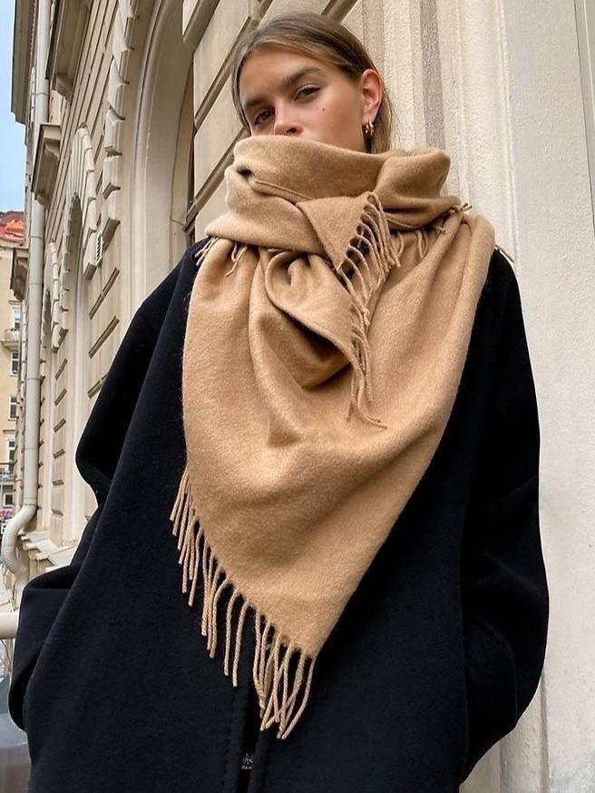 Person in a black coat and tan scarf with fringe stands against elegant architecture.