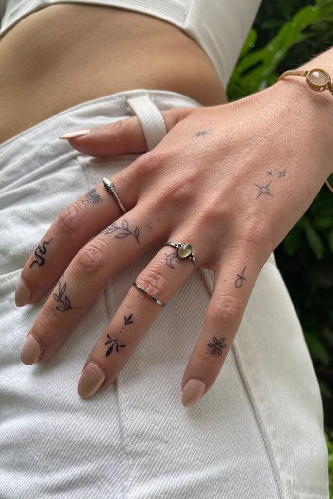 Close-up of a hand with minimalist finger tattoos and multiple rings, wearing a white outfit, surrounded by greenery.