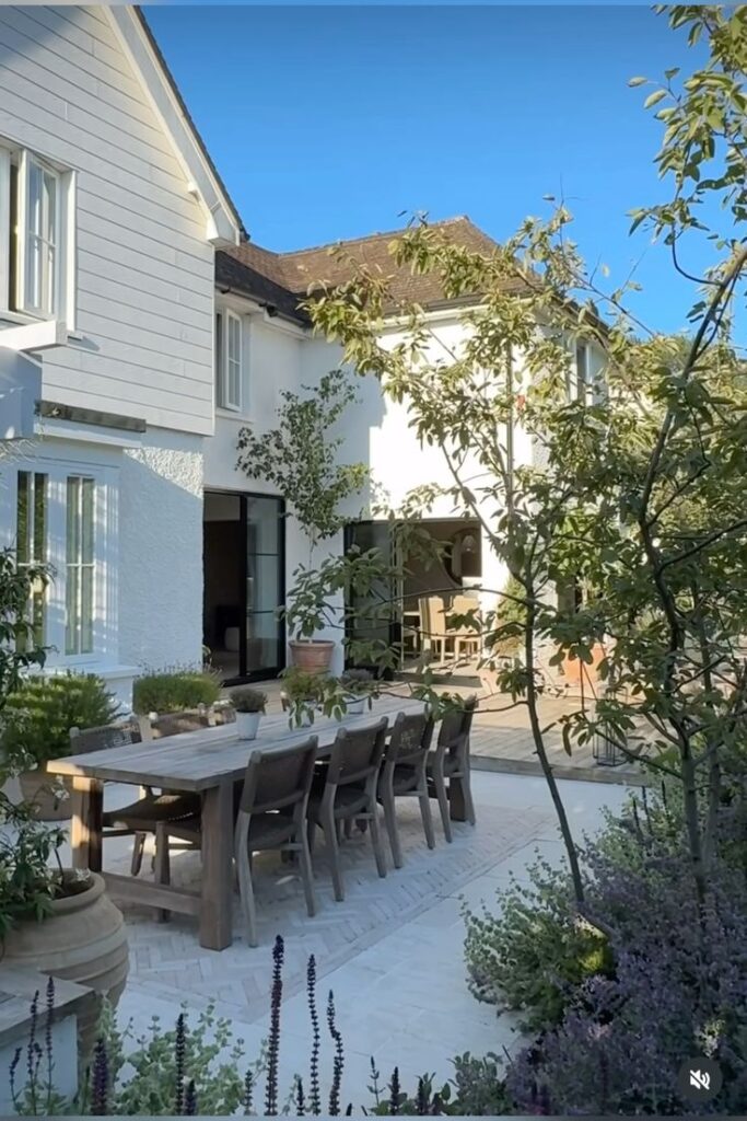 Elegant outdoor dining area with wooden table in a lush garden, beside a beautiful white house under a clear blue sky.