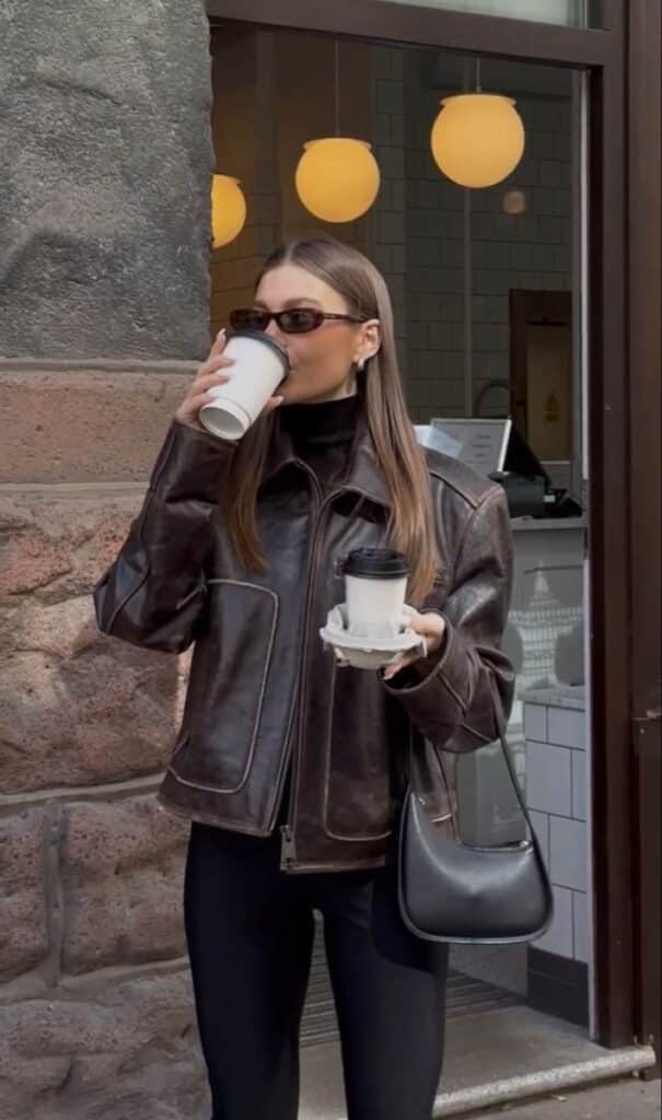 Stylish woman in sunglasses drinks coffee while holding two cups outside a cafe, wearing a leather jacket.