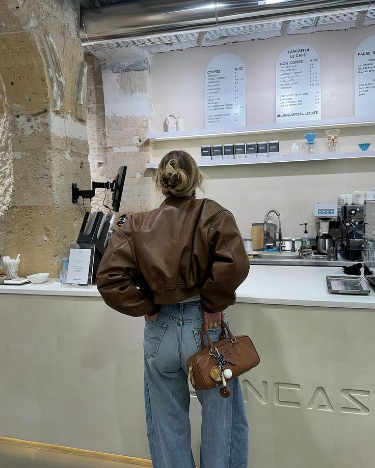 Woman in a cafe wearing a brown jacket and jeans, holding a handbag, with a menu on the wall.