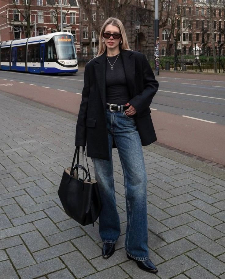 Fashionable woman in black blazer and jeans with tote bag stands on street near tram.