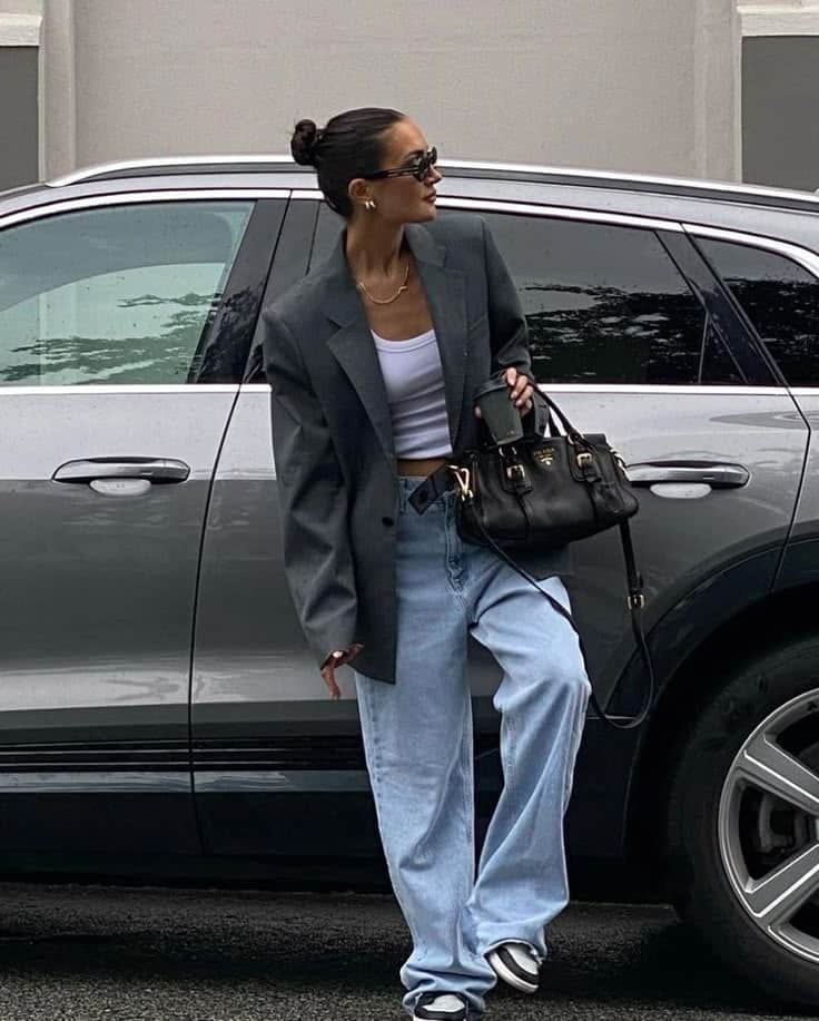 Woman in blazer and jeans poses by car, holding black handbag, fashion street style.