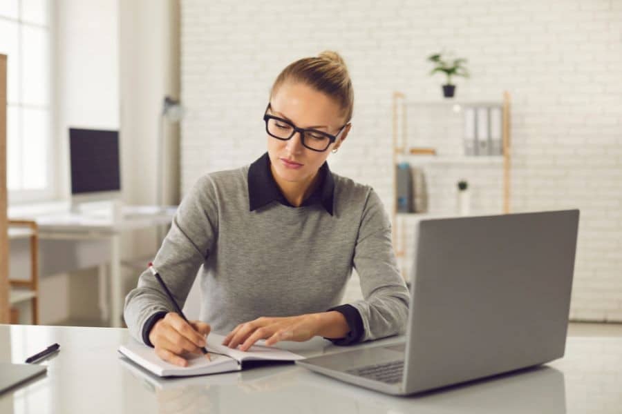 Professional woman with glasses writing in a notebook at her desk with a laptop in a modern office setting.