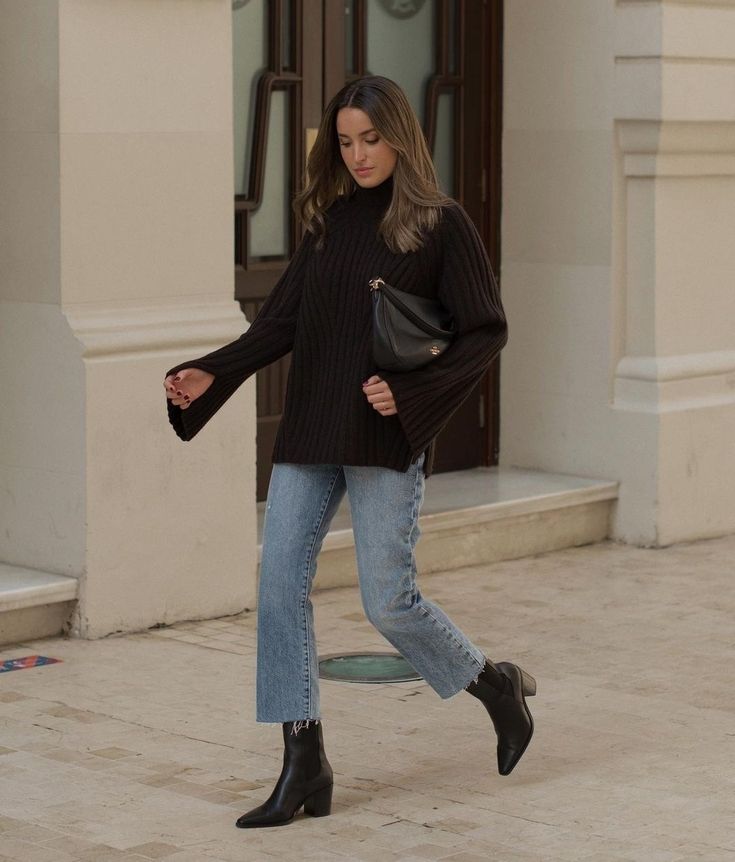 Woman in stylish black sweater and jeans walking confidently outdoors. Fashionable outfit with boots and handbag.