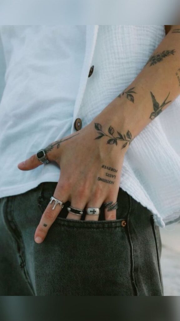 Hand with leaf tattoos and silver rings resting in pocket, wearing a casual white shirt and dark jeans.