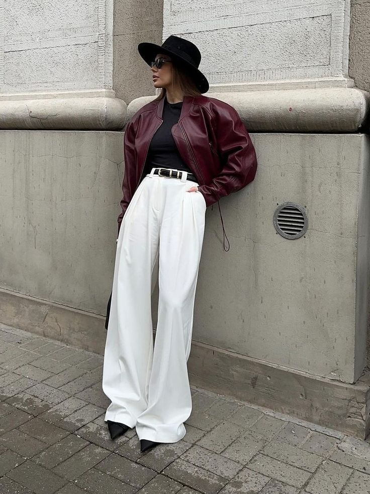 Stylish woman in a burgundy jacket, white pants, and wide-brimmed hat stands against an urban backdrop.