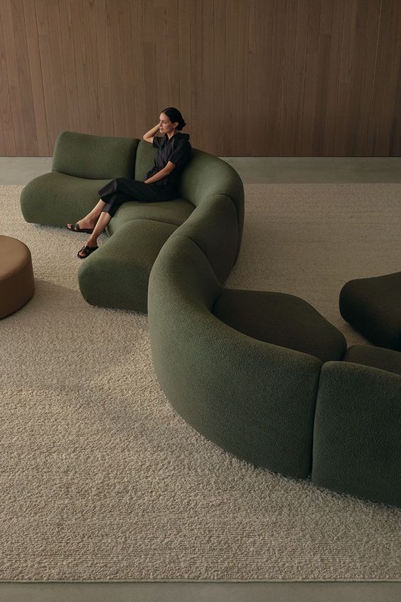 Modern living room with woman on curved, green sofa and beige carpet, wooden wall backdrop.