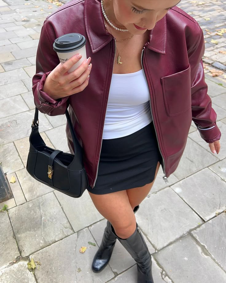 Woman in burgundy jacket with coffee cup and black bag, walking on paved street.