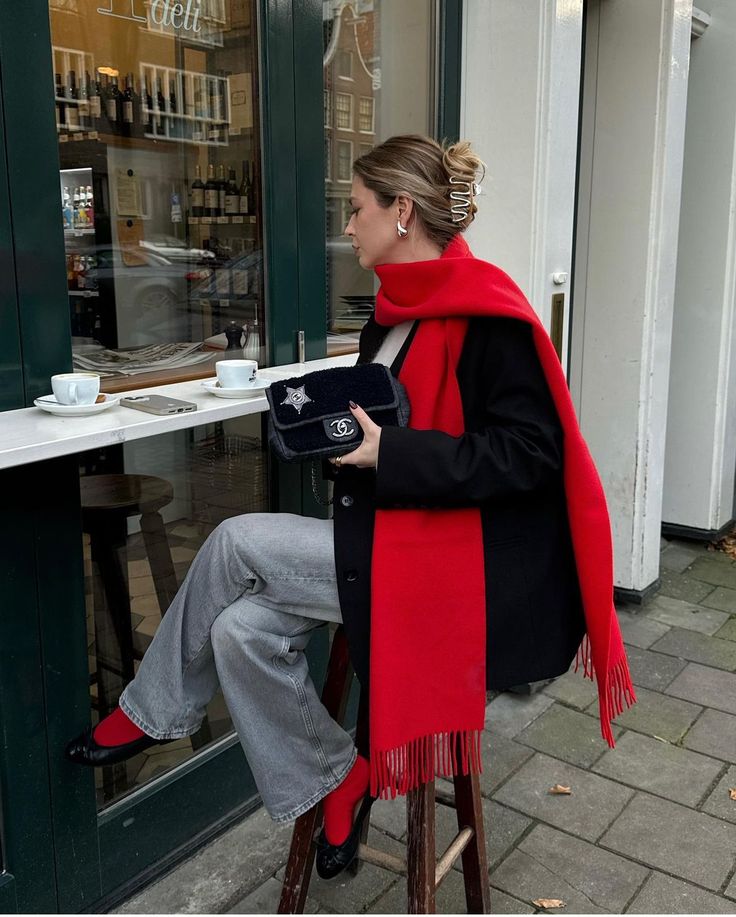 Woman in red scarf sits outside café, holding a bag, with coffee cups on the table and a stylish outfit.
