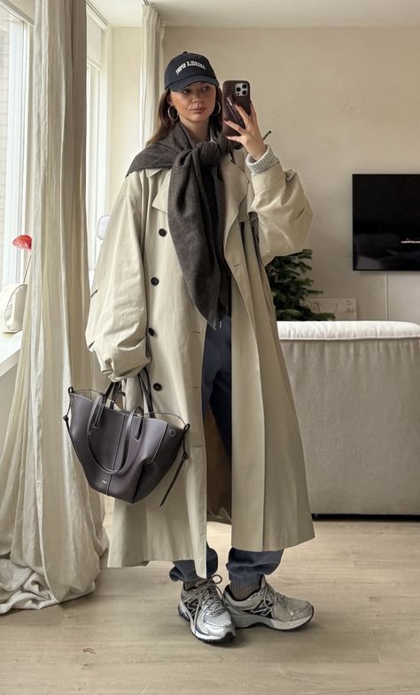 Fashionable woman in long coat with handbag and cap takes a mirror selfie in a stylish living room.