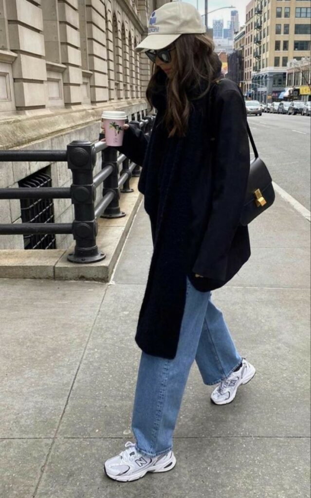 A woman in casual streetwear walks with a coffee cup in a city setting, wearing jeans, sneakers, and a baseball cap.