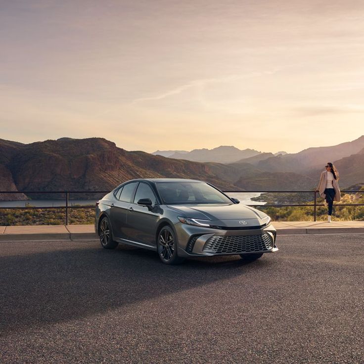 Sleek grey car parked by scenic mountain view at sunset, woman walking nearby, highlighting travel and adventure.