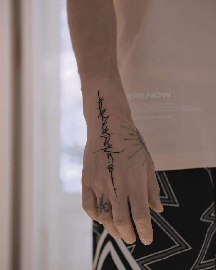 Close-up of a hand with intricate black tattoos, including a script design and sun-like patterns, on a light background.